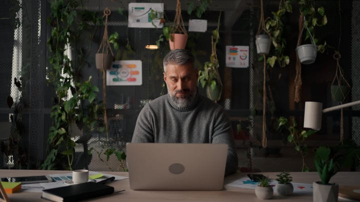 Senior Man Working On Laptop At Desk In Modern Green Office
