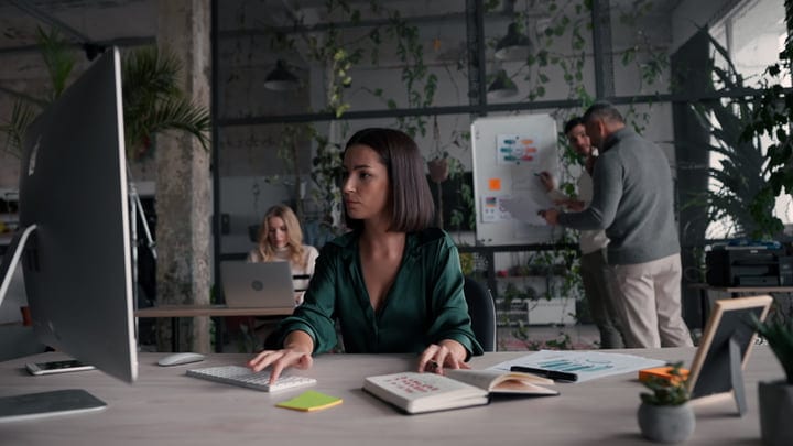 Focused Businesswoman Typing Amidst a Busy Office Environment