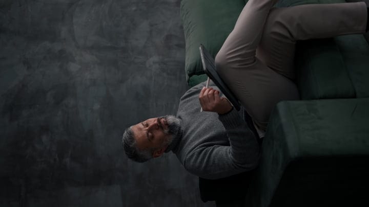 Mature Bearded Man Working Calmly On A Tablet While Lying On A Sofa