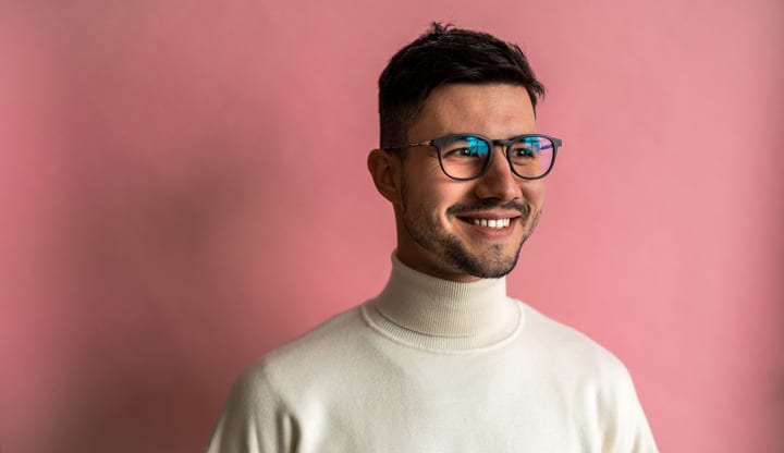 Young Man With Glasses Smiling Wearing White Turtleneck Against Pink Background