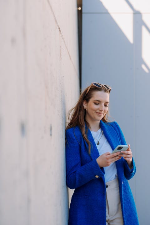 Young Woman in Blue Coat Texting by Sunlit Wall