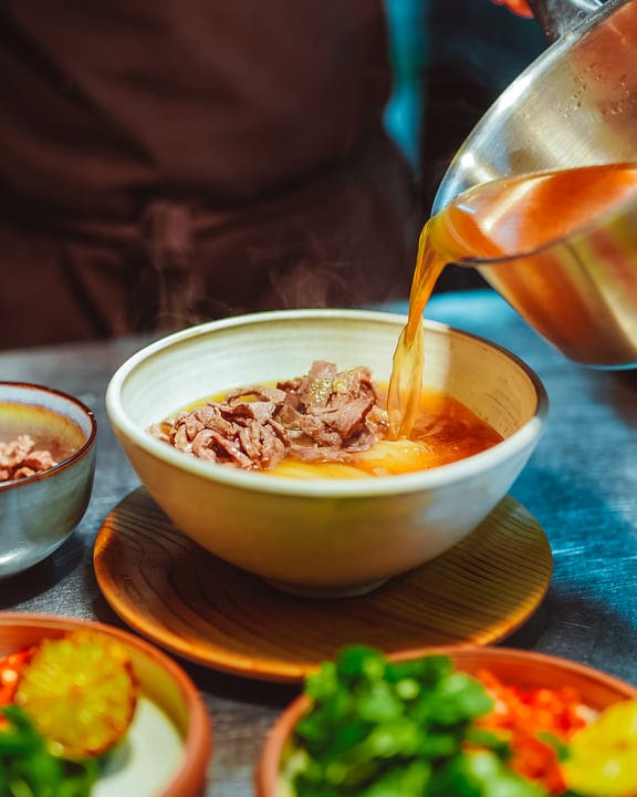 Pouring Rich Broth Into Hearty Beef Noodle Soup Bowl