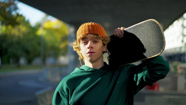 Young Man With Orange Beanie Carrying Skateboard On His Shoulder Outdoors