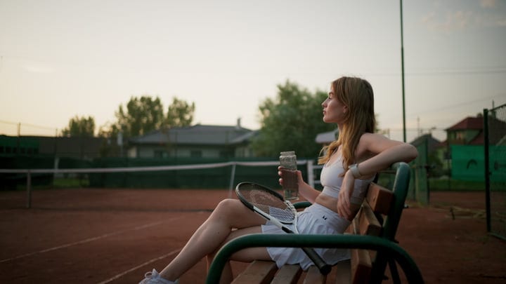 Female Tennis Player Relaxing On Court Bench With Drink At Sunset