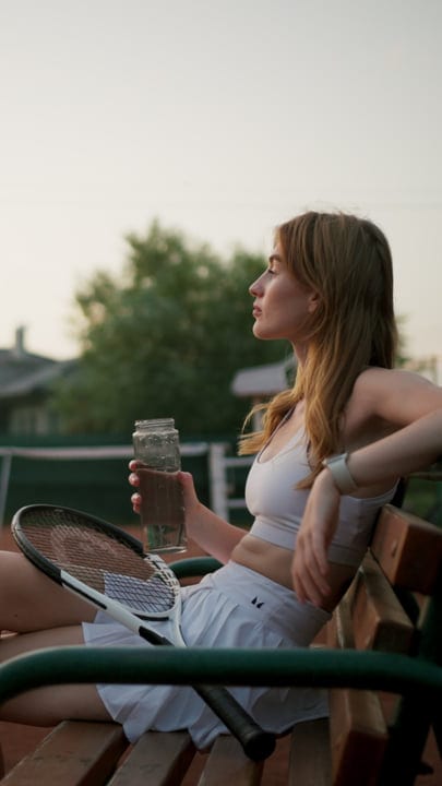 A Woman Tennis Player Rests On A Court Bench With Water