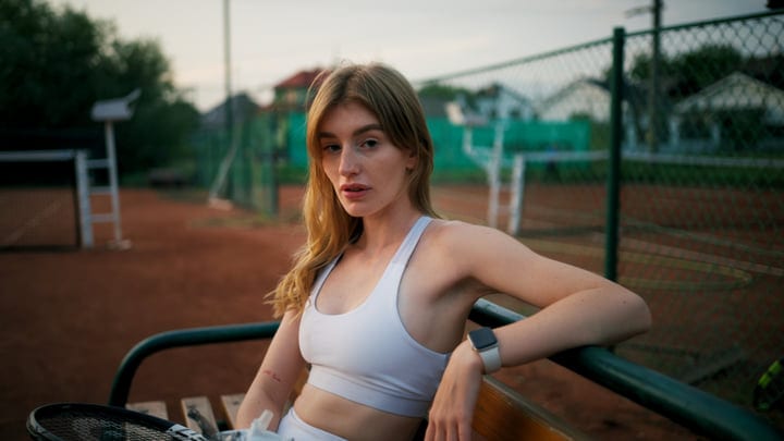 Young Woman Sits on Bench with Racket on Outdoor Tennis Court