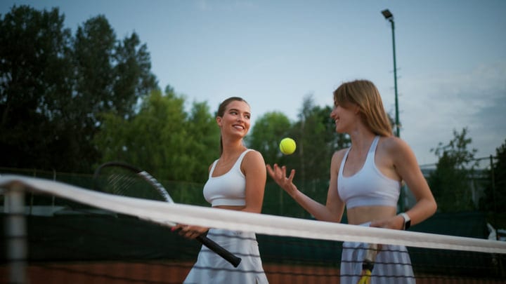 Two Female Athletes Tossing A Tennis Ball On An Outdoor Court