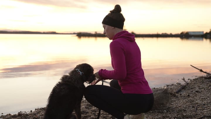 Woman Kneeling Beside Her Pet Dog On A Rocky Shore At Sunset