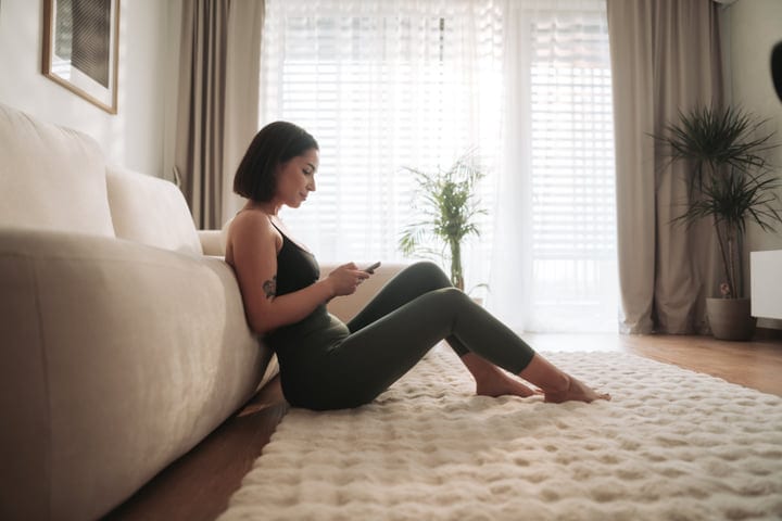 Woman In Activewear Comfortably Browsing Phone Next To Sofa In Her Apartment
