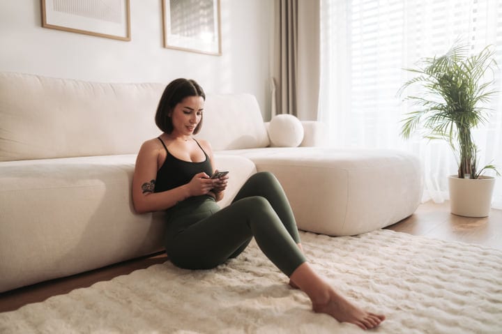 Young European Woman Sits On A Fluffy Rug Using Her Phone At Home