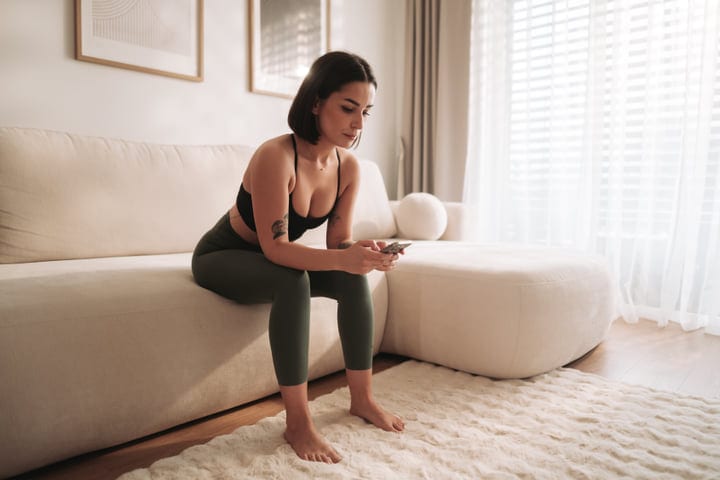 Young Woman Sitting On Sofa Checking Smartphone In Bright Modern Living Room