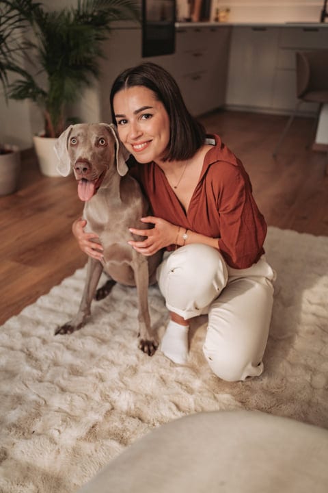 Young Woman Kneeling And Hugging Her Weimaraner Dog On A Rug Indoors