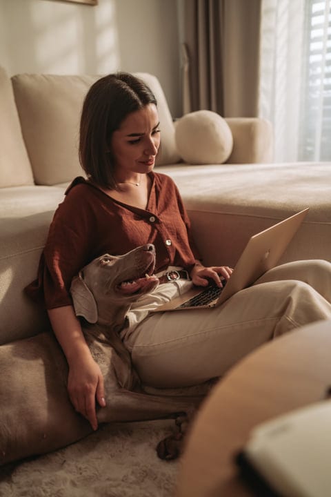 Woman Working On Laptop With Her Dog Relaxing Beside Her On Couch