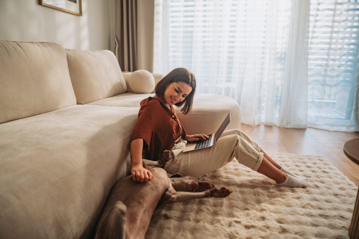 Smiling Woman Sits On Floor With Her Laptop, Petting Dog By Sofa