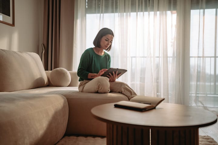 Young Woman Sitting On Couch Using Digital Tablet In Bright Living Room