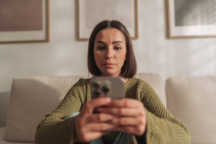 Young Woman Sitting Comfortably On Sofa Using Her Smartphone In A Bright Living Room