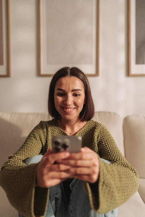 Young Woman Smiles While Checking Her Smartphone On A Comfortable Sofa At Home