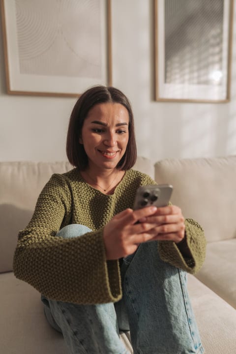 Young Woman Smiling And Using Her Mobile Phone On A Sofa