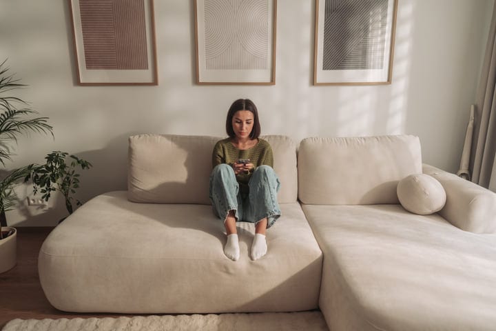 Young Woman Sits On Modern Sofa Browsing Smartphone In Bright European Apartment