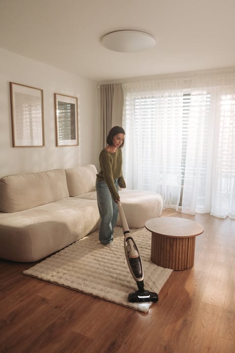 Young Woman Cleans Living Room Rug With Cordless Vacuum On Bright Morning