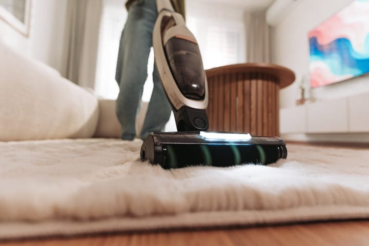 A Woman Vacuuming A Light Shaggy Rug With A Cordless Cleaner