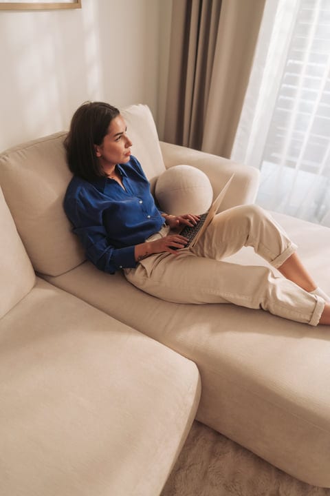 Woman Lying On Sofa With Laptop Looking Out Window In Bright Room