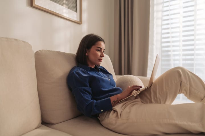 Young Woman Typing On Laptop While Reclining On A Sofa At Home
