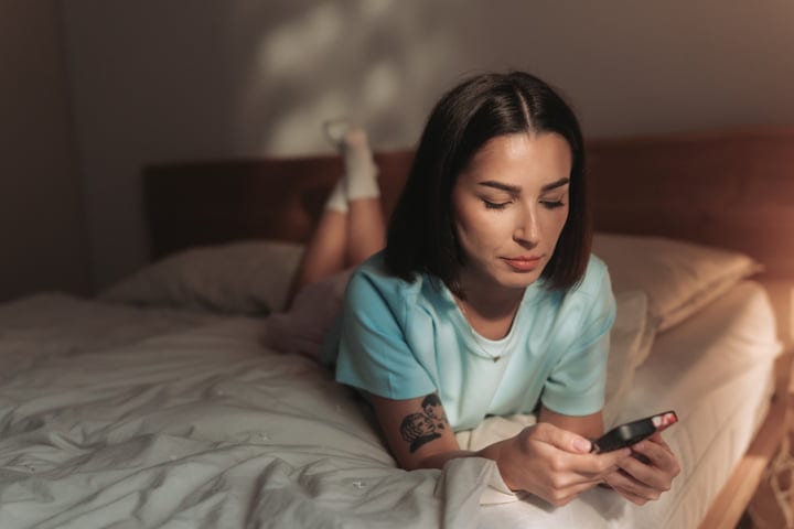 Young Woman Lying On Bed Browsing Social Media On Smartphone