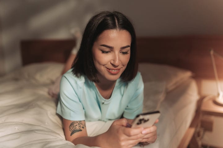 Young Woman Lying On Bed Browsing Social Media On Her Phone In Bedroom