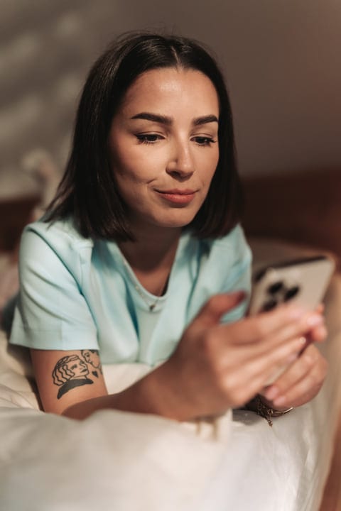 Young Woman Relaxing In Bed Scrolling On Her Smartphone Morning Routine