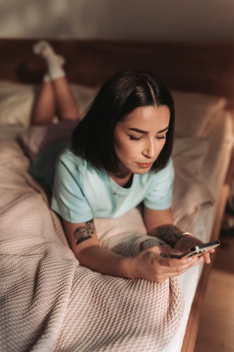 Young Woman Lying On Bed Checking Phone In Cozy Morning Light