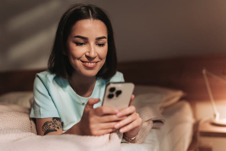 Young Woman Lying In Bed And Smiling While Using Her Smartphone