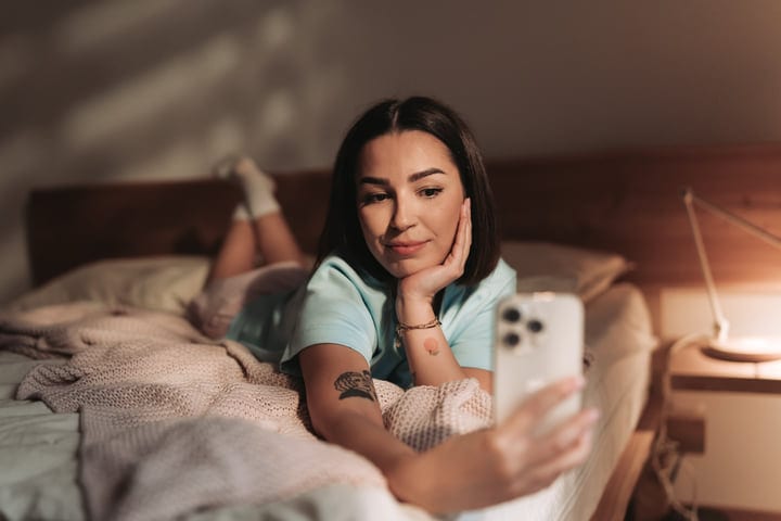 Young European Woman Lying On Bed Using Smartphone In Her Bedroom