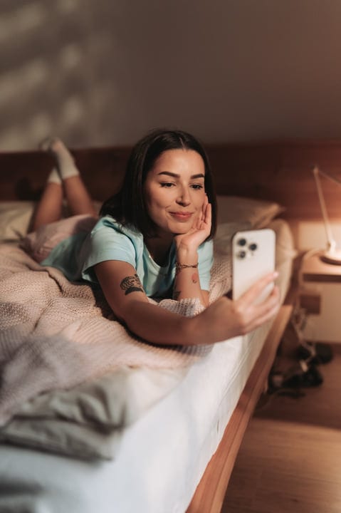 Young Woman Lying On Bed Browsing Phone In Sunny Bedroom