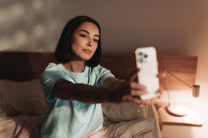 Young Woman Holding Smartphone Taking Selfie In Her Cozy Bedroom