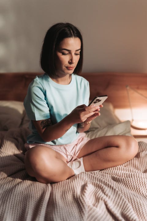 Young Woman Cross-Legged On Bed Checking Smartphone In Soft Morning Light