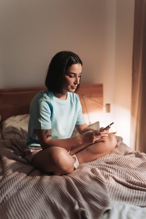Young Woman Checking Her Mobile Phone While Sitting On Bed In Warm Morning Light