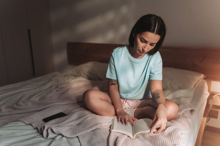 Young Woman Sits Comfortably On Bed Reading A Book In The Early Morning