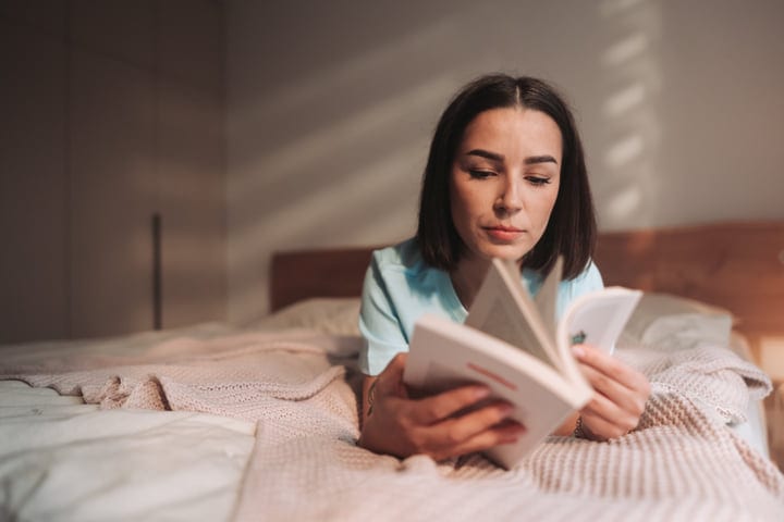 Focused Woman Reads Book Lying On Her Bed In Soft Bedroom Light