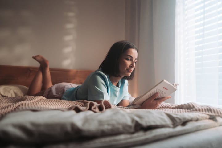 Young Woman Relaxing In Bed Reading A Book By The Window At Home