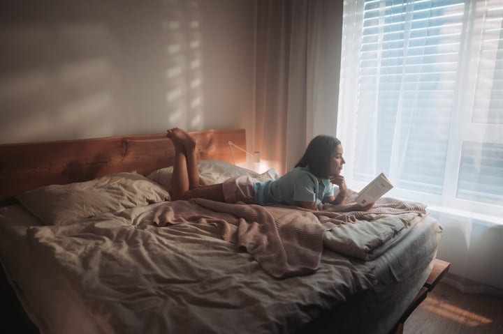 Young Woman Reading A Book In Bed By Window During Morning Light