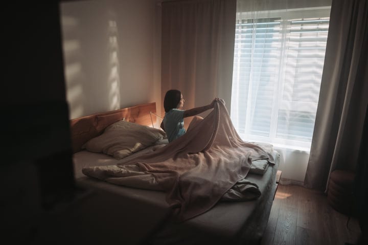 A Woman Sits In Bed Holding Blanket Gazing At Bright Window
