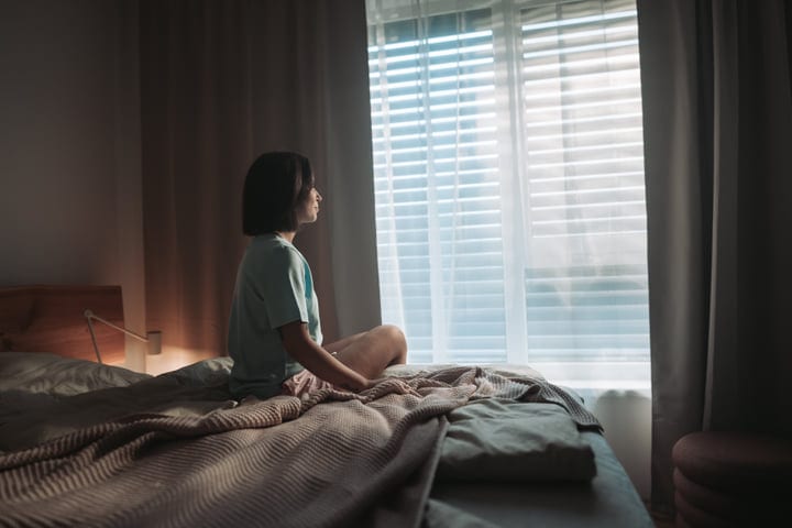 Woman Sits On Her Bed Looking Out Window Into Bright Morning Light
