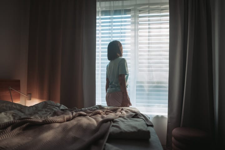 Woman Standing By Window In Bedroom During Early Morning