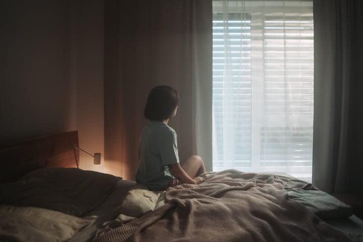 Woman Sits On Bed Looking Out Window Through Blinds In Morning Light