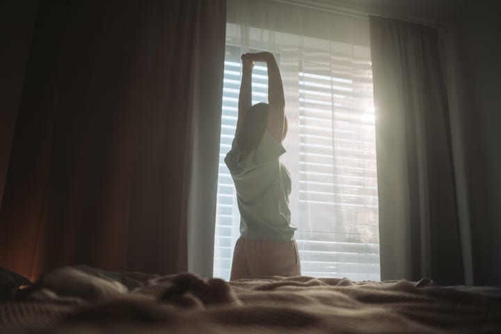 Woman Stretches By Window In Bedroom Bathed In Bright Morning Sunlight