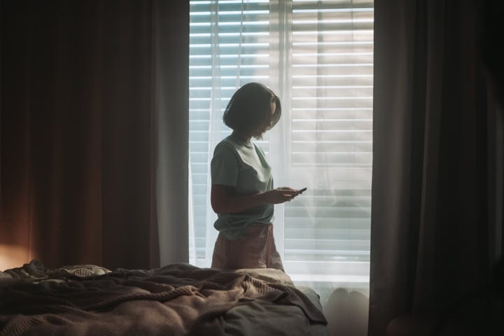 Young Woman Checking Phone By Window In Bedroom Morning Light