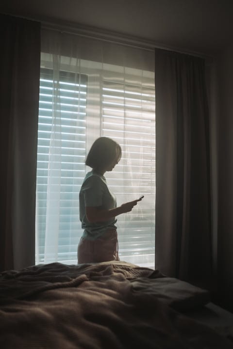 Woman Stands Silhouetted By Bedroom Window Checking Phone In Morning Light