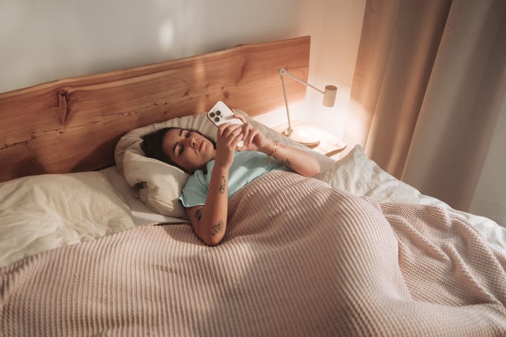 Young Woman Lying In Bed Browsing On Her Smartphone In The Morning