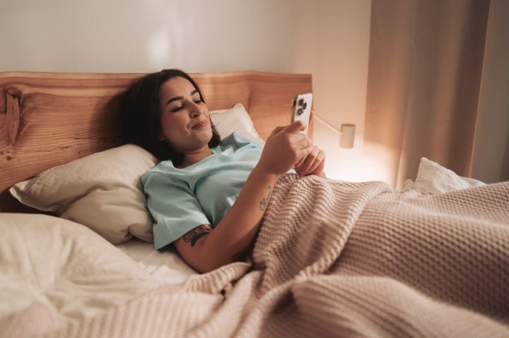 Young Woman Smiling Browsing Smartphone In Cozy Bed During Morning